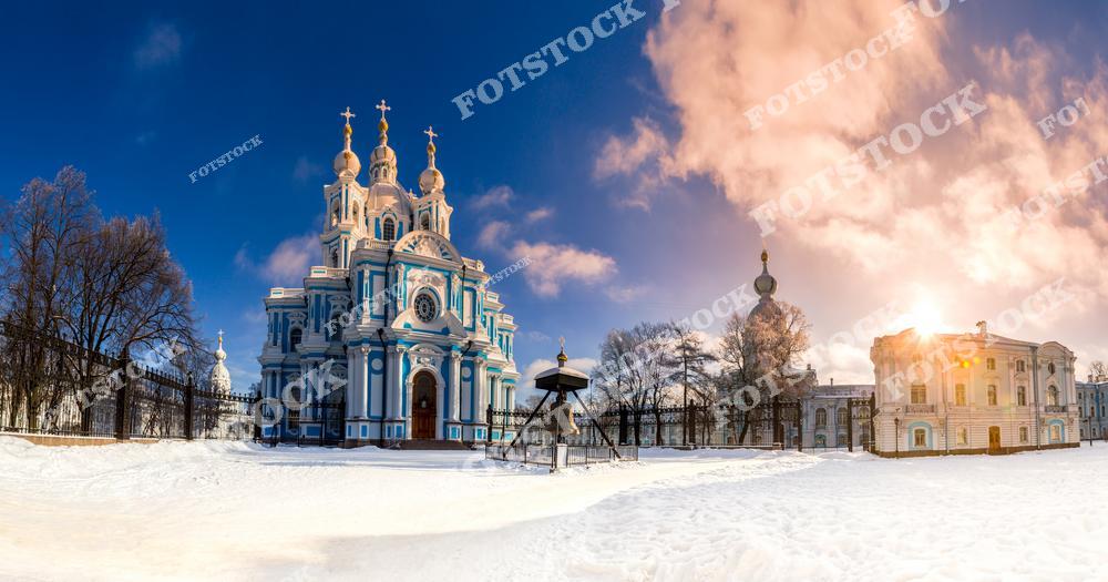 Фотография зимнего Санкт-Петербурга с панорамным видом на центр города и величественный Смольный собор. На снимке запечатлены исторические улицы, музеи и архитектурные памятники, а заснеженные крыши и набережные Невы создают атмосферу зимней сказки северной столицы России. Зимний свет мягко освещает фасады зданий, подчеркивая богатое культурное наследие Петербурга. Этот кадр прекрасно подходит для туристических публикаций, коллекций зимних фотографий, постеров, оформления интерьеров и сувенирной продукции, демонстрируя красоту и уникальность зимнего Петербурга.